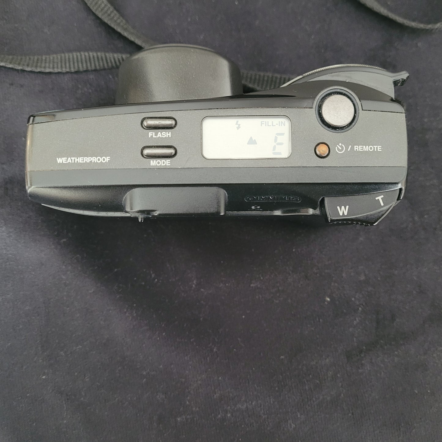 Draufansicht der Olympus Superzoom 120 Multi AF Analogkamera in Schwarz, mit sichtbarem Schriftzug Superzoom 120 auf der Oberseite. Das herausfahrbare Zoomobjektiv Olympus Lens Zoom 35-120mm ist gut zu erkennen. Die Kamera liegt auf einer dunklen Oberfläche, mit Trageriemen an der Seite – ein perfektes Vintage-Fotografie-Highlight für Analogkamera-Liebhaber.