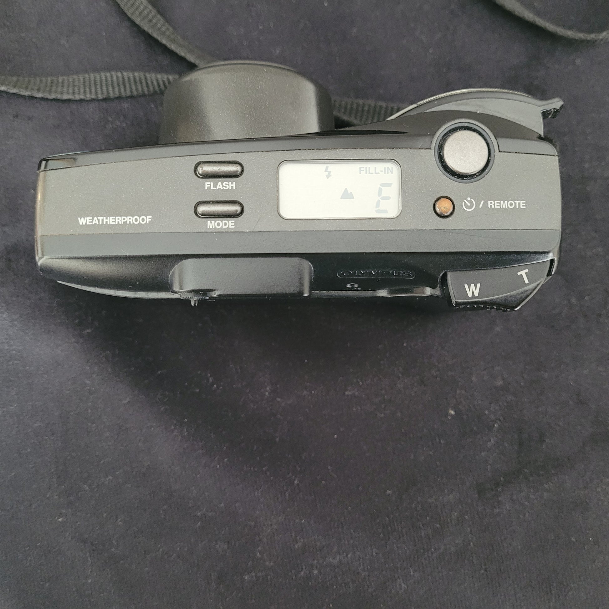 Draufansicht der Olympus Superzoom 120 Multi AF Analogkamera in Schwarz, mit sichtbarem Schriftzug Superzoom 120 auf der Oberseite. Das herausfahrbare Zoomobjektiv Olympus Lens Zoom 35-120mm ist gut zu erkennen. Die Kamera liegt auf einer dunklen Oberfläche, mit Trageriemen an der Seite – ein perfektes Vintage-Fotografie-Highlight für Analogkamera-Liebhaber.