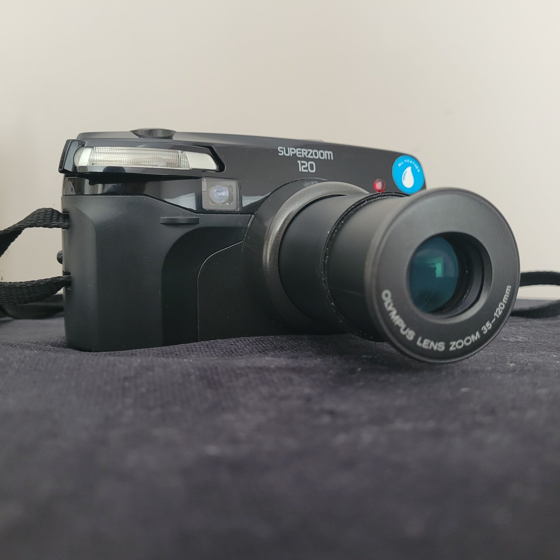 Olympus Superzoom 120 Multi AF Analogkamera – klassische Vintage Analogkamera mit ausgefahrenem 35-120mm Olympus Zoomobjektiv. Die elegante schwarze Kamera liegt auf dunklem Stoff und zeigt deutlich die Modellbezeichnung Superzoom 120. Perfekt für Liebhaber hochwertiger Retro-Fotografie.