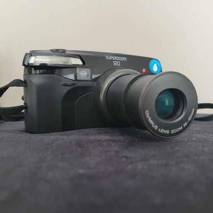 Olympus Superzoom 120 Multi AF Analogkamera – klassische Vintage Analogkamera mit ausgefahrenem 35-120mm Olympus Zoomobjektiv. Die elegante schwarze Kamera liegt auf dunklem Stoff und zeigt deutlich die Modellbezeichnung Superzoom 120. Perfekt für Liebhaber hochwertiger Retro-Fotografie.