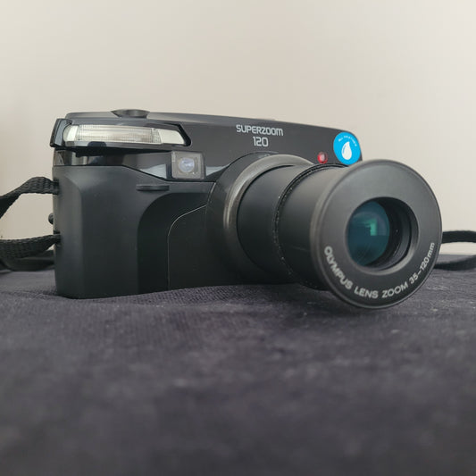 Olympus Superzoom 120 Multi AF Analogkamera – klassische Vintage Analogkamera mit ausgefahrenem 35-120mm Olympus Zoomobjektiv. Die elegante schwarze Kamera liegt auf dunklem Stoff und zeigt deutlich die Modellbezeichnung Superzoom 120. Perfekt für Liebhaber hochwertiger Retro-Fotografie.