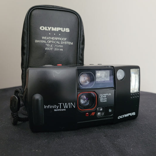 Vintage Olympus AF-1 Infinity Twin Quartzdate Kamera mit 35-mm- und 70-mm-Objektiv, ausgestattet mit Biaxial Optical System. Wetterfeste Kamera in Schwarz mit roten Akzenten, geprüft ohne Film und funktionstüchtig. Neben der Kamera eine schwarze Olympus-Tasche mit Modellaufdruck, ideal für Liebhaber der analogen Fotografie und Sammler von Vintage-Kameras.