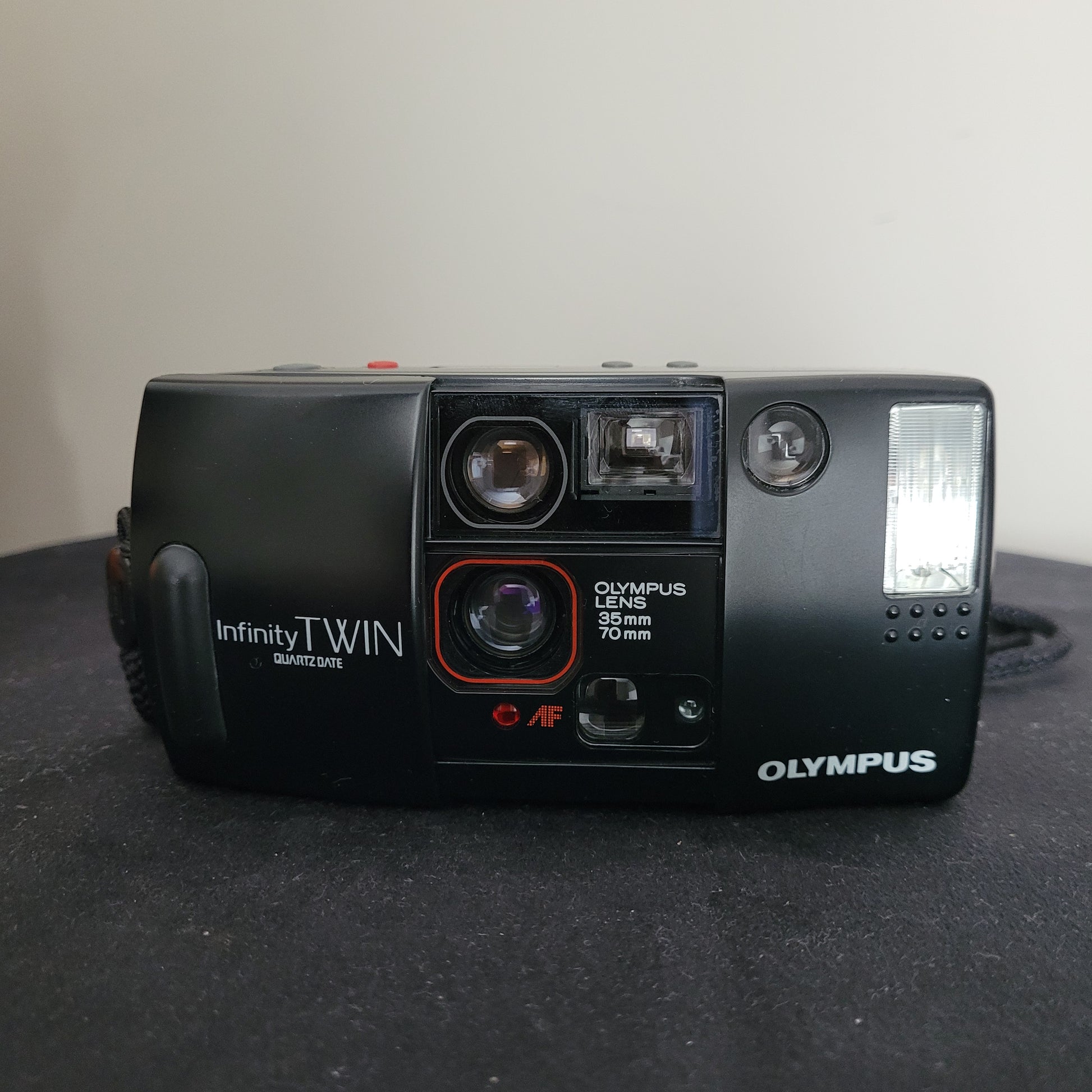 Vintage Olympus AF-1 Infinity Twin Quartzdate Kamera mit wetterfestem Design, zwei integrierten Objektiven (35 mm Weitwinkel und 70 mm Teleobjektiv) und Quartzdate-Funktion. Gezeigt neben ihrer schwarzen Originalschutztasche mit Modellaufdruck. Die Kamera wurde ohne Film geprüft und ist voll funktionstüchtig. Ideal für Sammler von Vintage-Kameras und Liebhaber der analogen Fotografie.