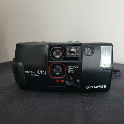 Vintage Olympus AF-1 Infinity Twin Quartzdate Kamera mit wetterfestem Design, zwei integrierten Objektiven (35 mm Weitwinkel und 70 mm Teleobjektiv) und Quartzdate-Funktion. Gezeigt neben ihrer schwarzen Originalschutztasche mit Modellaufdruck. Die Kamera wurde ohne Film geprüft und ist voll funktionstüchtig. Ideal für Sammler von Vintage-Kameras und Liebhaber der analogen Fotografie.