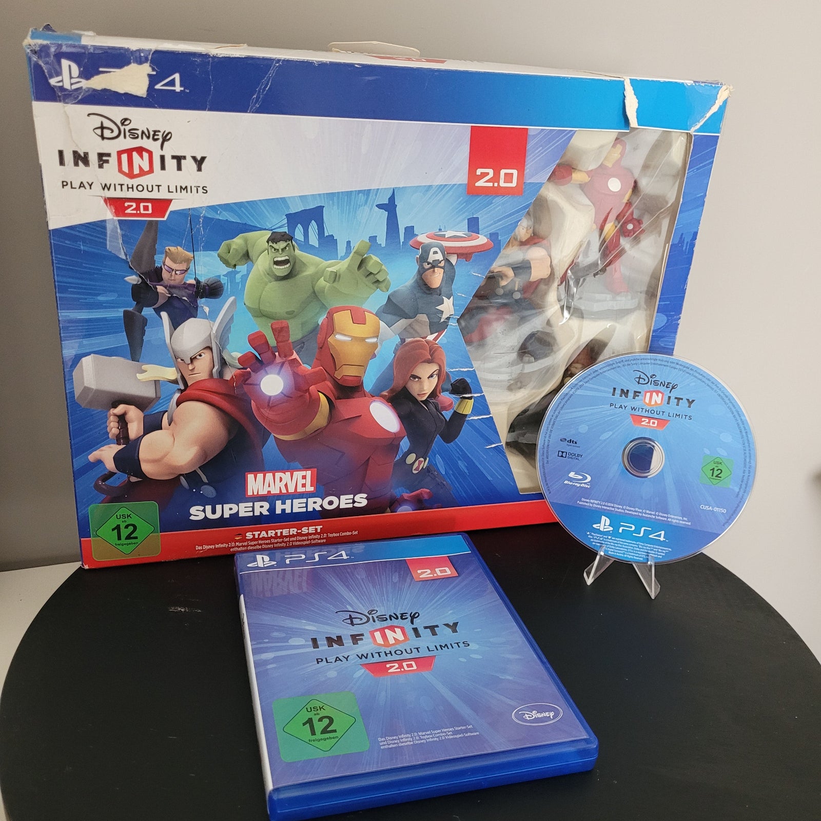 Die Vorderseite des PS4 Disney Infinity 2.0 Marvel Super Heroes Startersets mit der passenden Spiel-CD. Das Set liegt auf einem schwarzen Untergrund, während der Hintergrund weiß ist.