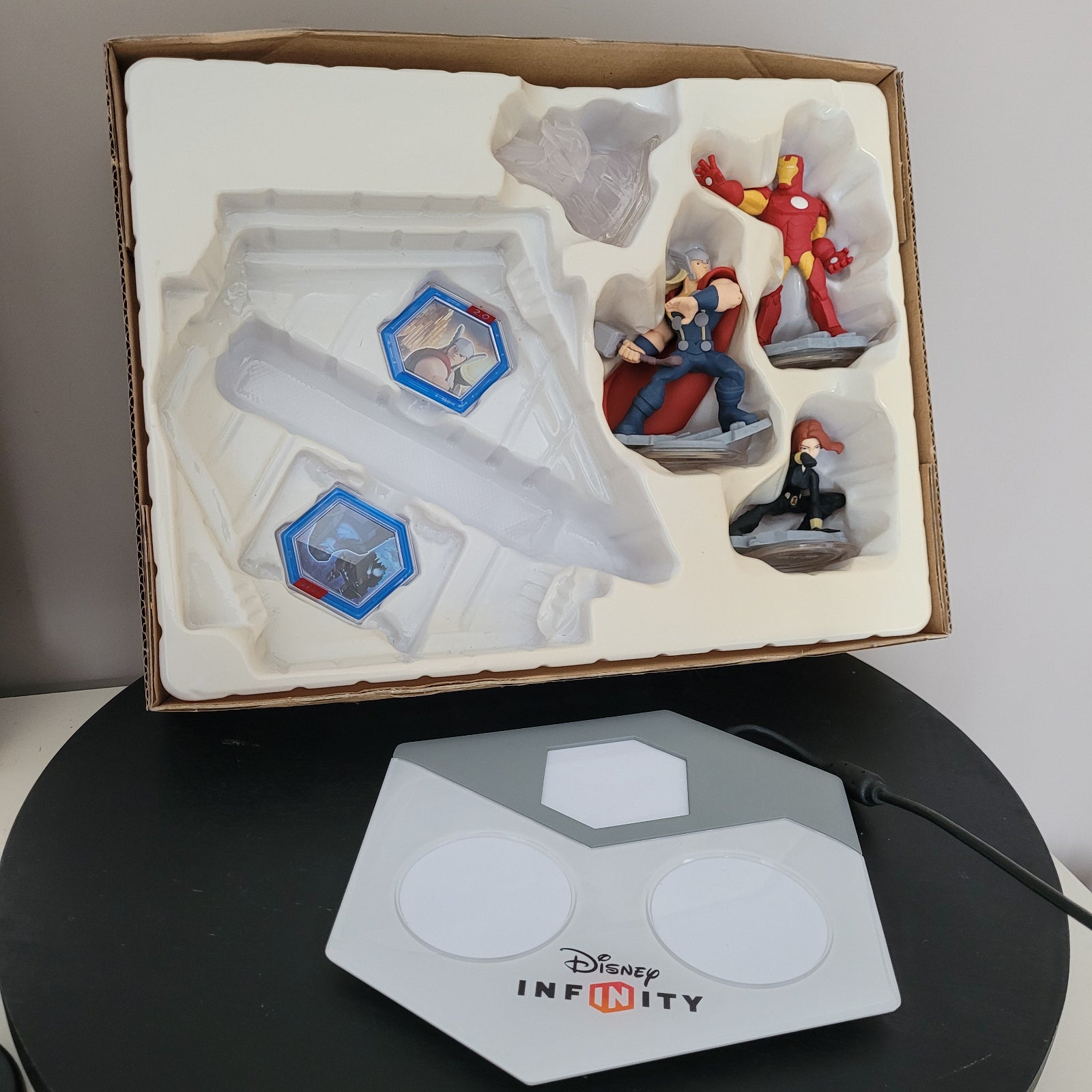 Das Innere der Verpackung des PS4 Disney Infinity 2.0 Marvel Super Heroes Startersets ist sichtbar. Im Vordergrund befindet sich das Disney Infinity-Portal, auf dem die Figuren platziert werden. Die Verpackung zeigt verschiedene Fächer für die Spiel-CD und die dazugehörigen Sammelfiguren. Die Elemente sind übersichtlich angeordnet, wodurch das Set eine strukturierte Präsentation erhält.