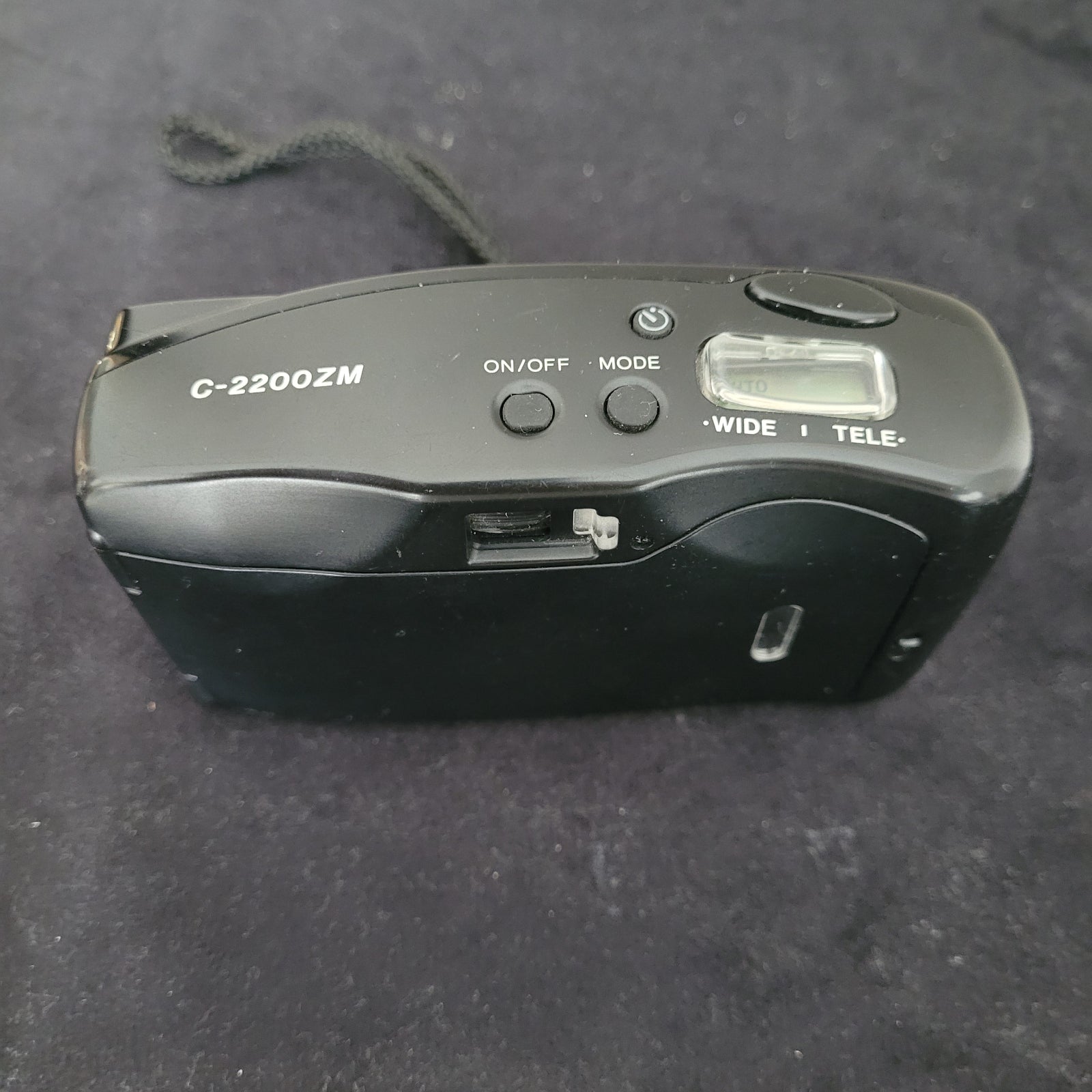 Das Bild zeigt die Draufsicht der Vintage Panasonic Analogkamera, platziert auf einem schwarzen Untergrund mit weißem Hintergrund. Sichtbar sind die Bedienelemente der Kamera, darunter der Auslöser, verschiedene Einstellungsräder sowie der integrierte Blitz, der sich auf der rechten Seite befindet.