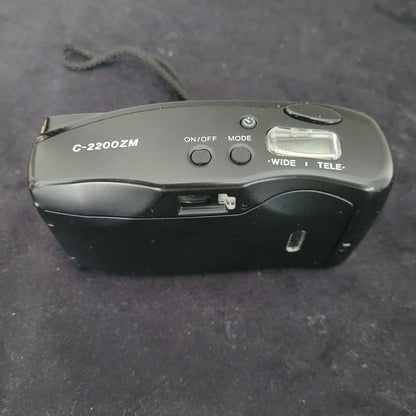 Das Bild zeigt die Draufsicht der Vintage Panasonic Analogkamera, platziert auf einem schwarzen Untergrund mit weißem Hintergrund. Sichtbar sind die Bedienelemente der Kamera, darunter der Auslöser, verschiedene Einstellungsräder sowie der integrierte Blitz, der sich auf der rechten Seite befindet.