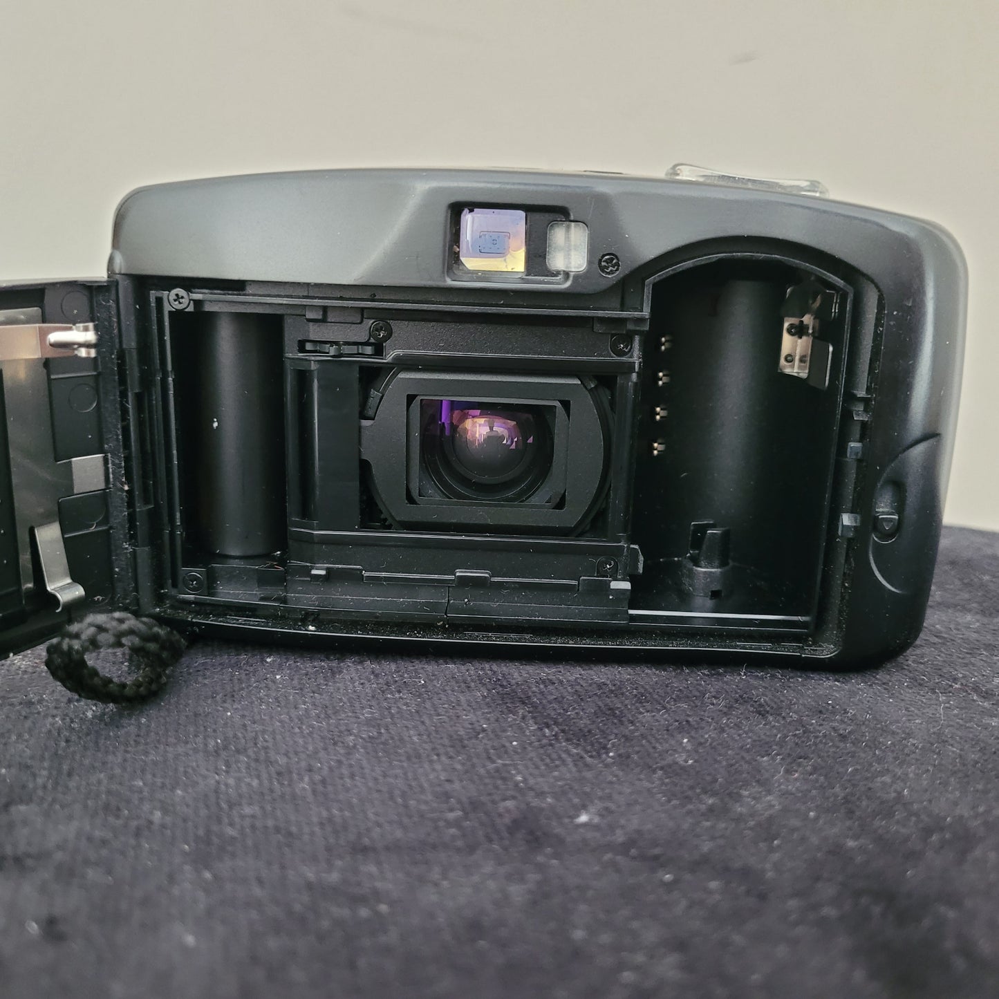 Das Bild zeigt das geöffnete Filmfach der Vintage Panasonic Analogkamera, platziert auf einem schwarzen Untergrund mit weißem Hintergrund. Sichtbar sind die Filmaufnahmespule, die Führungselemente für den Filmtransport sowie die Kameramechanik, die für analoge Aufnahmen notwendig ist.