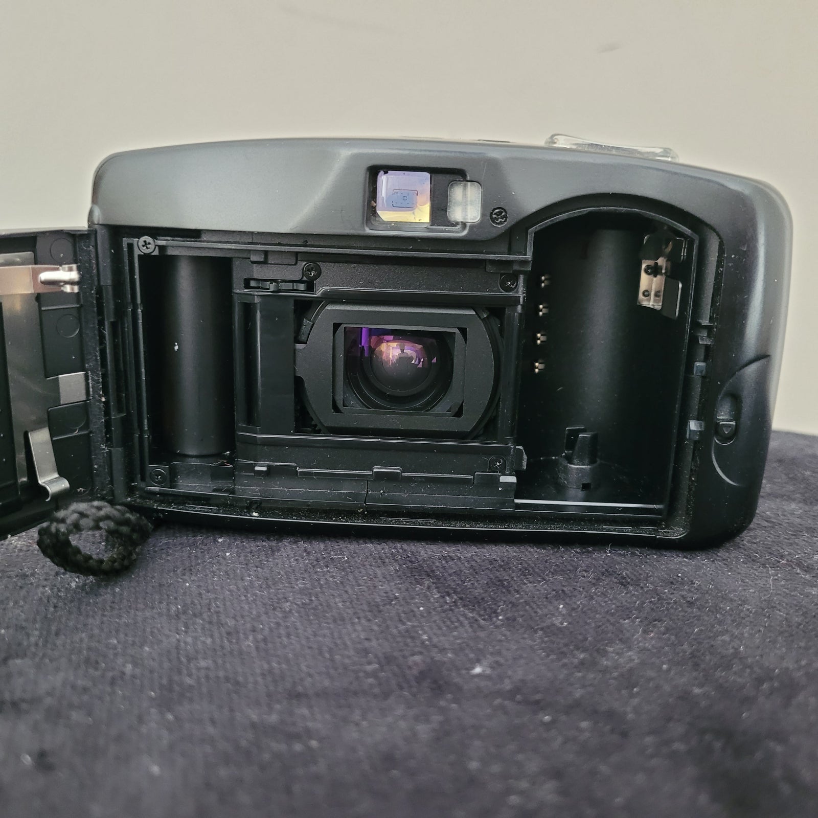 Das Bild zeigt das geöffnete Filmfach der Vintage Panasonic Analogkamera, platziert auf einem schwarzen Untergrund mit weißem Hintergrund. Sichtbar sind die Filmaufnahmespule, die Führungselemente für den Filmtransport sowie die Kameramechanik, die für analoge Aufnahmen notwendig ist.