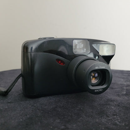 Das Bild zeigt die Seitenansicht der Vintage Panasonic Analogkamera mit Zoom-Funktion, platziert auf einem schwarzen Untergrund. Der weiße Hintergrund hebt das kompakte Design sowie die 35-70mm Macro-Objektivbeschriftung und den integrierten Blitz hervor.