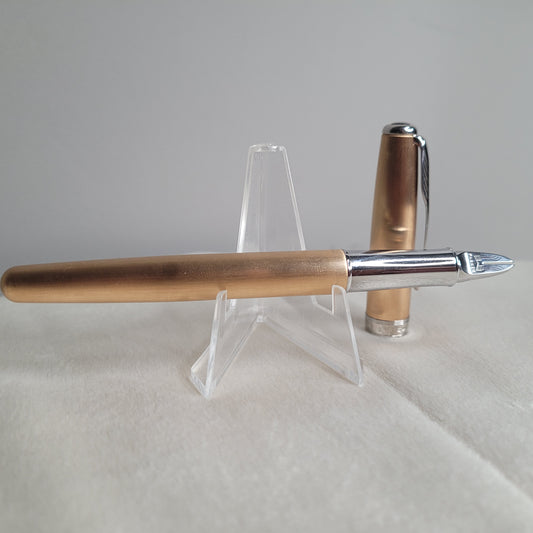 Das Bild zeigt einen Parker Füllfederhalter mit der 5th Technologie in der Ausführung Sonnet Rosé Gold & Chrome. Der Füllfederhalter liegt auf einem durchsichtigen Ständer, wobei die Kappe des Stiftes daneben liegt. Der Stift hat eine roségoldene Oberfläche mit einem glänzenden Chromgriff und einer verzierten Feder. Die Kappe ist ebenfalls in Roségold gehalten und hat einen silbernen Clip.