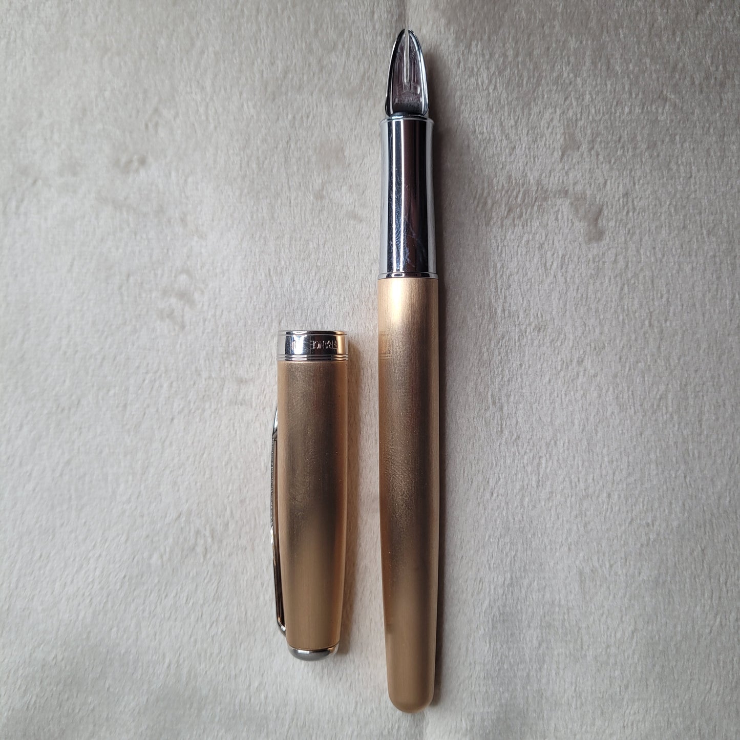 Das Bild zeigt einen Parker Füllfederhalter mit der 5th Technologie aus der Sonnet Serie in Rosé Gold und Chrom. Der Füllfederhalter liegt auf einer hellen, weichen Oberfläche. Der Stift ist in zwei Teile zerlegt: die Kappe und der Hauptkörper des Füllfederhalters. Die Kappe ist aus Rosé Gold mit einem silbernen Clip und einem silbernen Ring am unteren Ende, auf dem \"PARKER\" eingraviert ist. Der Hauptkörper des Füllfederhalters ist ebenfalls in Rosé Gold gehalten, mit einem silbernen Abschnitt