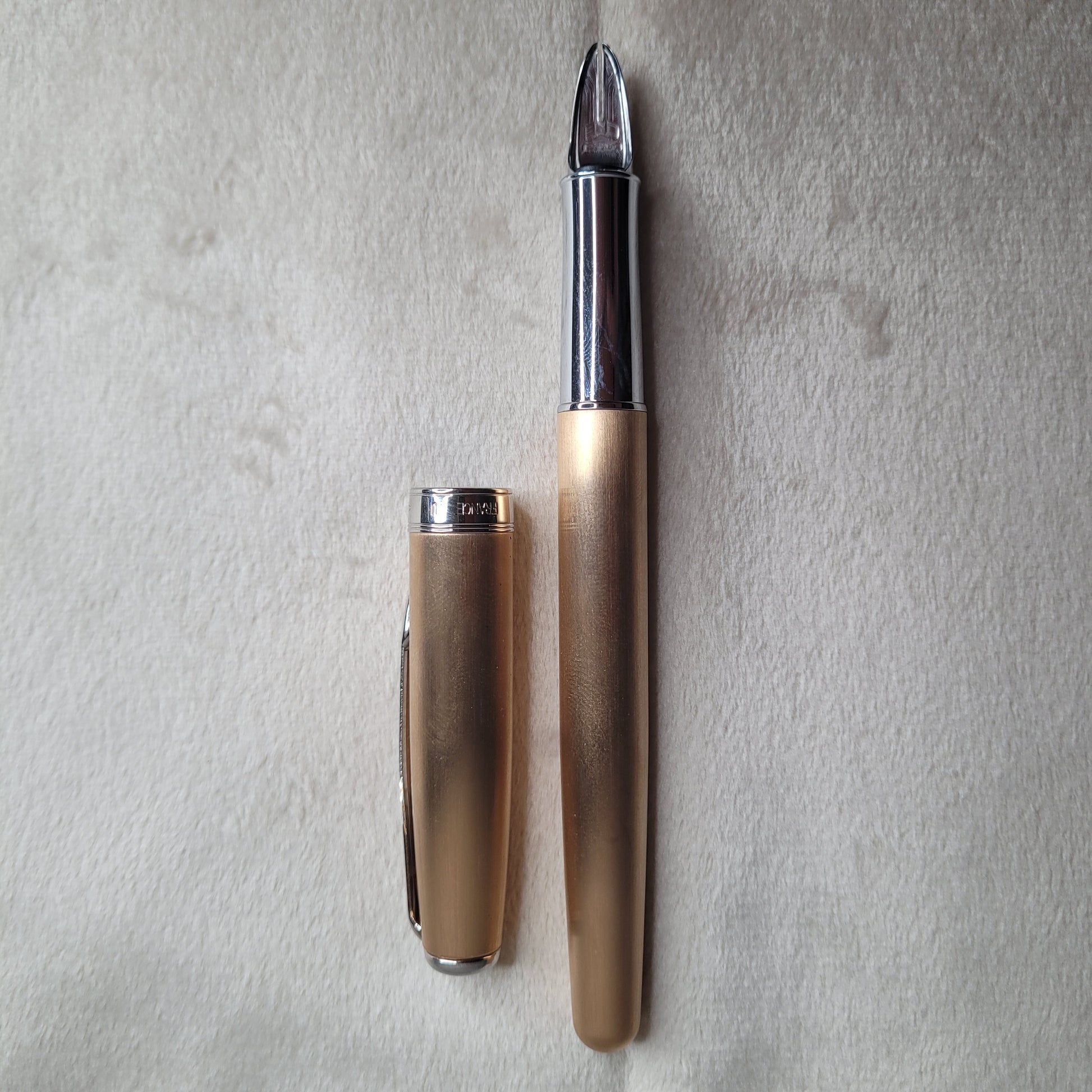 Das Bild zeigt einen Parker Füllfederhalter mit der 5th Technologie aus der Sonnet Serie in Rosé Gold und Chrom. Der Füllfederhalter liegt auf einer hellen, weichen Oberfläche. Der Stift ist in zwei Teile zerlegt: die Kappe und der Hauptkörper des Füllfederhalters. Die Kappe ist aus Rosé Gold mit einem silbernen Clip und einem silbernen Ring am unteren Ende, auf dem \"PARKER\" eingraviert ist. Der Hauptkörper des Füllfederhalters ist ebenfalls in Rosé Gold gehalten, mit einem silbernen Abschnitt