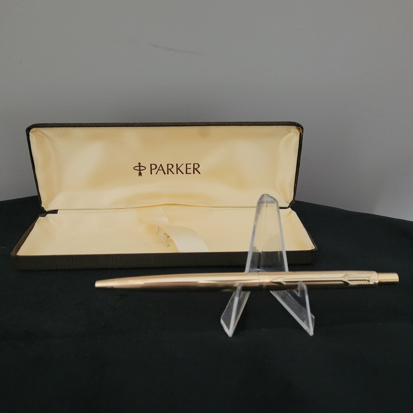 Ein goldfarbener Parker-Kugelschreiber in einer offenen, cremefarbenen Schachtel mit schwarzem Rand, auf einem schwarzen Hintergrund. Der Kugelschreiber steht auf einem transparenten Ständer.