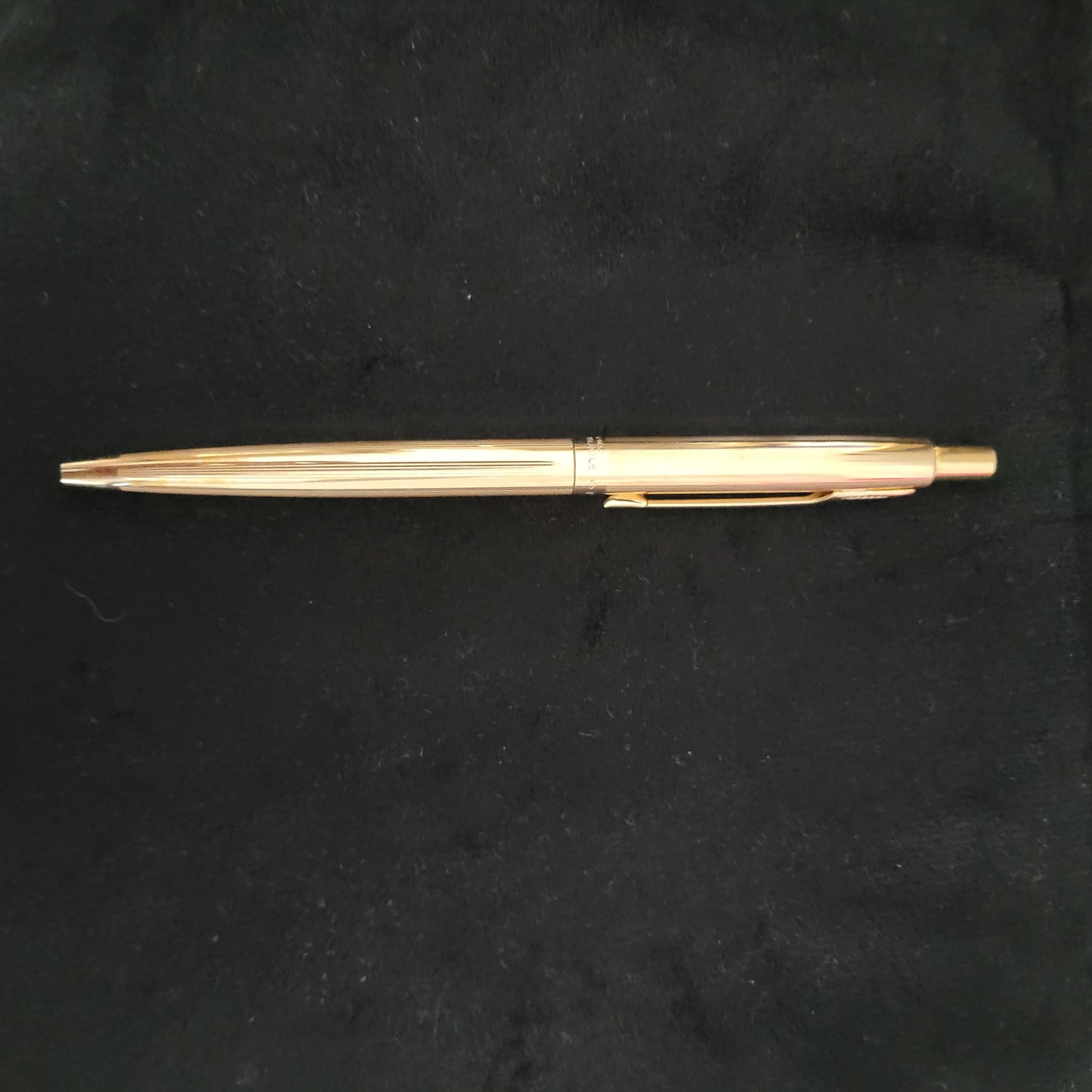 Ein goldfarbener Parker-Kugelschreiber liegt horizontal auf einem schwarzen Hintergrund. Der Stift hat eine glänzende Oberfläche mit einem Clip auf der oberen Seite und einem Druckknopfmechanismus am oberen Ende. Der Stiftkörper ist mit vertikalen Rillen verziert, die zu seiner ästhetischen Anziehungskraft beitragen.