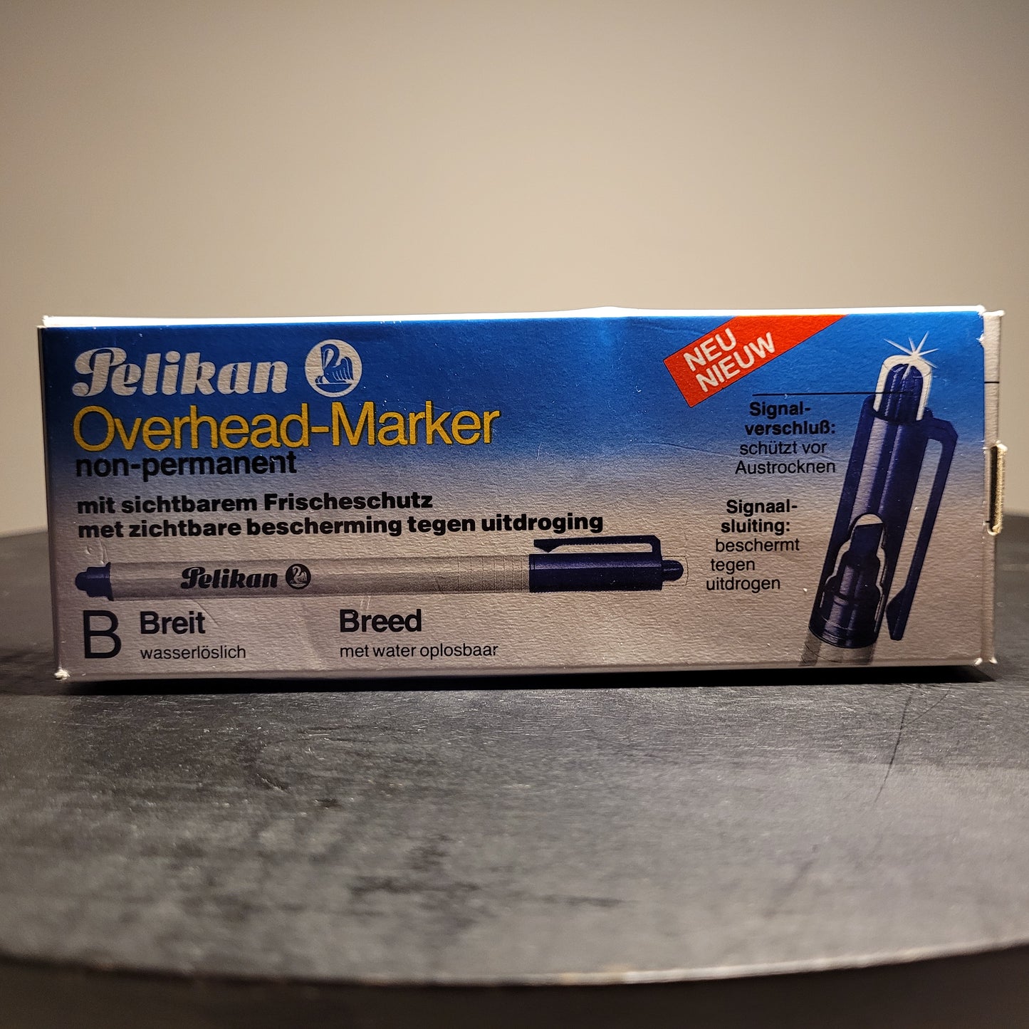 Die Abbildung zeigt eine Verpackung eines Pelikan Overhead-Markers. Der Marker ist nicht permanent und wasserlöslich. Auf der Verpackung steht „Pelikan Overhead-Marker non-permanent“ und „mit sichtbarem Frischeschutz“ sowie „mit zichtbare bescherming tegen uitdroging“. Es gibt auch einen Hinweis „NEU“ in der oberen rechten Ecke. Der Marker hat eine breite Spitze („B Breit“ und „Breed“) und ist wasserlöslich („wasserlöslich“ und „met water oplosbaar“).