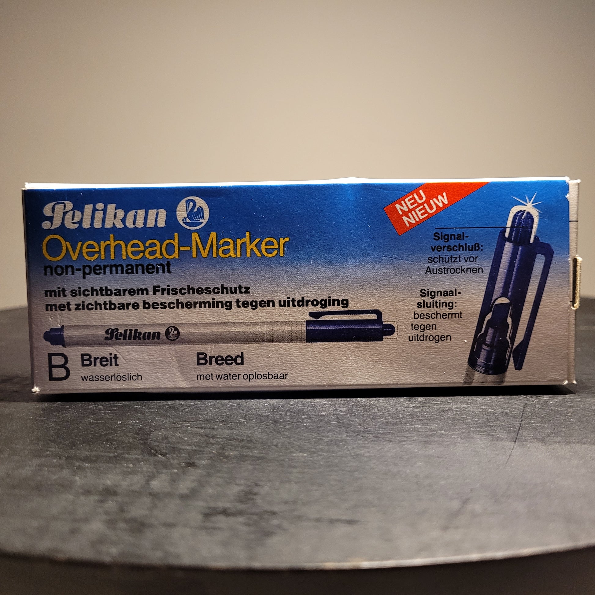 Die Abbildung zeigt eine Verpackung eines Pelikan Overhead-Markers. Der Marker ist nicht permanent und wasserlöslich. Auf der Verpackung steht „Pelikan Overhead-Marker non-permanent“ und „mit sichtbarem Frischeschutz“ sowie „mit zichtbare bescherming tegen uitdroging“. Es gibt auch einen Hinweis „NEU“ in der oberen rechten Ecke. Der Marker hat eine breite Spitze („B Breit“ und „Breed“) und ist wasserlöslich („wasserlöslich“ und „met water oplosbaar“).