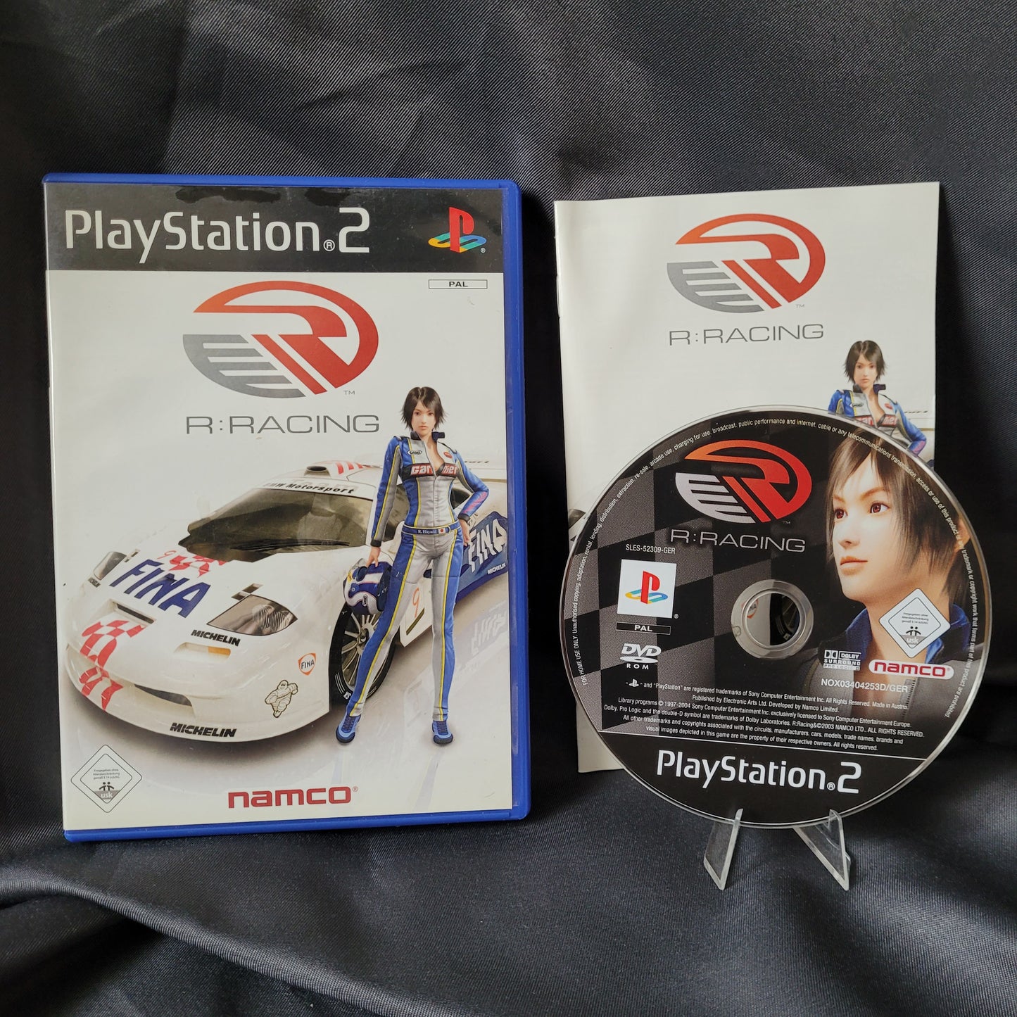 PlayStation 2 – PS2 – Spiel R:Racing Evolution von Namco (EAN 5030932037091) im Vintage-Sammlerzustand mit Spielhülle, Anleitung und Disc. Auf der Hülle ist eine Rennfahrerin neben einem weißen Rennwagen mit Sponsorenlogos abgebildet, Disc und Handbuch zeigen das gleiche Artwork mit Logo und Charakter.