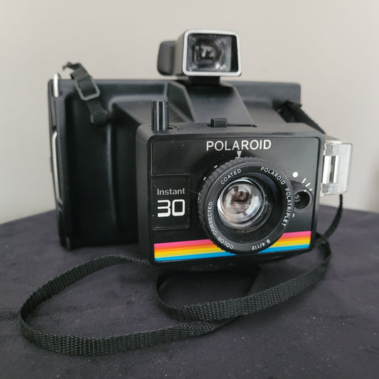 Polaroid Sofortbildkamera Modell Instant 30, schwarzes Gehäuse mit Regenbogenstreifen, aufgesetztem Sucher und Trageriemen. Ikonisches Design für Sammler und Fotografie-Enthusiasten.
