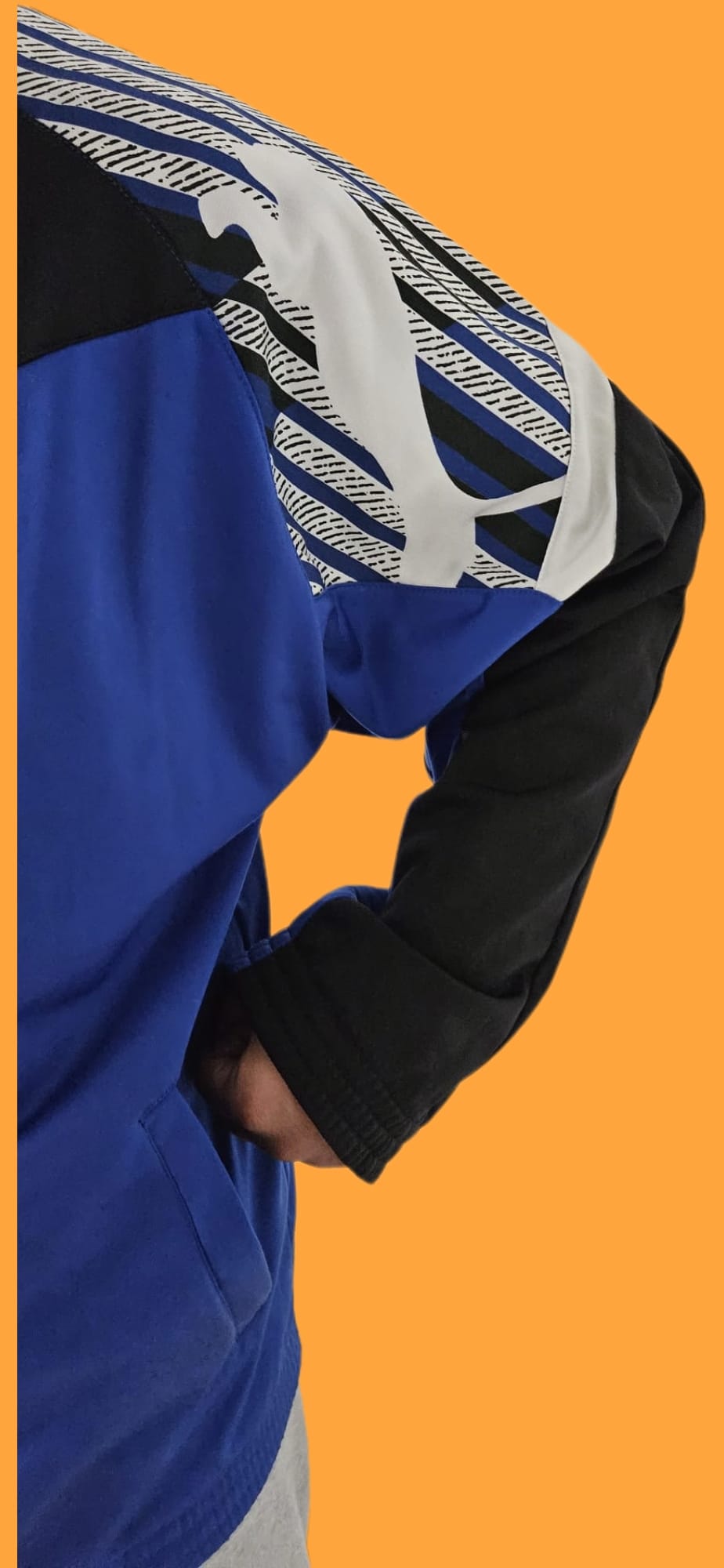Blau-schwarze Jacke mit weiß-schwarzem Muster am Ärmel, eine Hand lässig in der Tasche – urbaner Streetwear-Stil vor orangefarbenem Hintergrund.