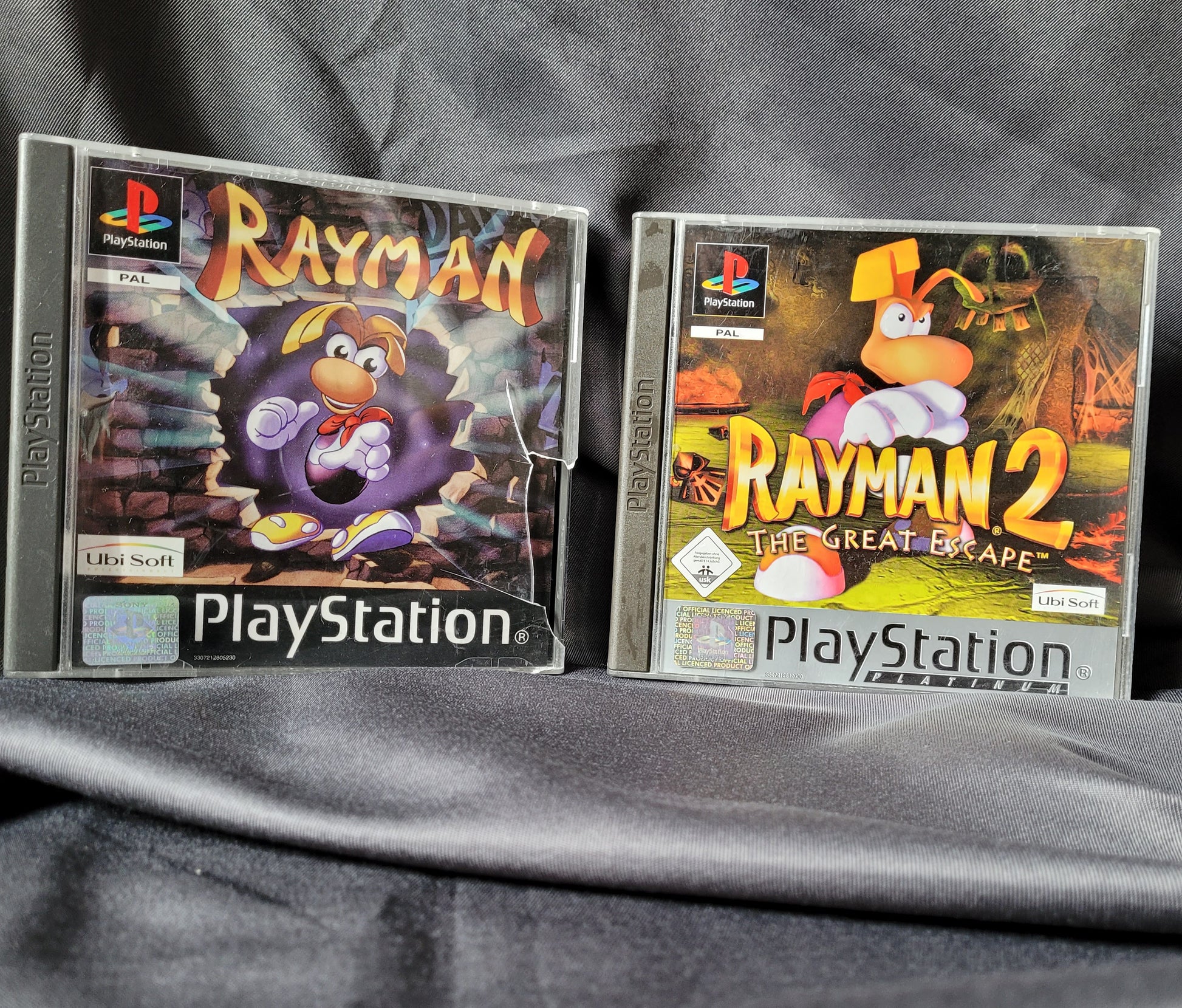 Zwei PlayStation 1 Spielehüllen auf dunklem Stoff: links Rayman mit bunter Höhlen-Grafik und Rayman-Figur, rechts Rayman 2 The Great Escape mit dynamischer Pose vor feurigem Hintergrund. Beide Spiele im PAL-Format mit PlayStation-Logo und Ubisoft-Branding.