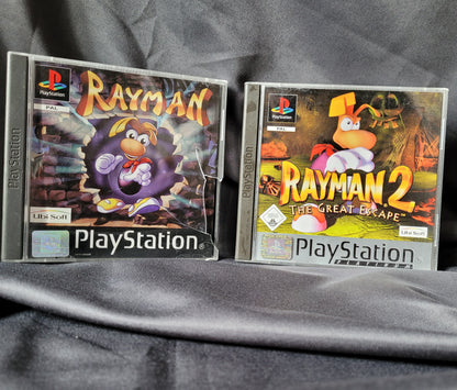 Zwei PlayStation 1 Spielehüllen auf dunklem Stoff: links Rayman mit bunter Höhlen-Grafik und Rayman-Figur, rechts Rayman 2 The Great Escape mit dynamischer Pose vor feurigem Hintergrund. Beide Spiele im PAL-Format mit PlayStation-Logo und Ubisoft-Branding.