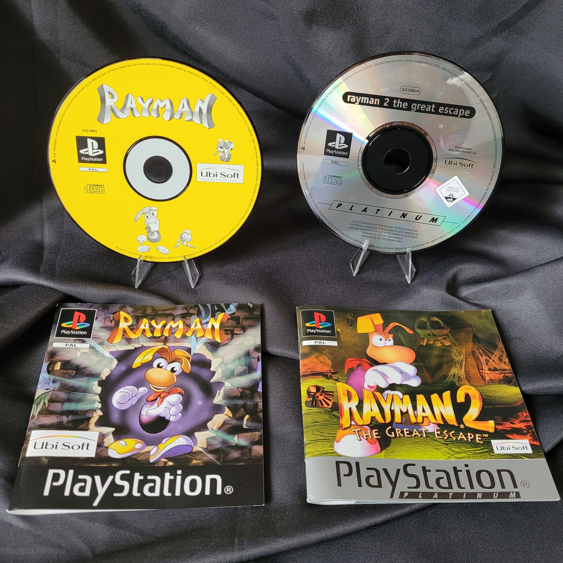 Zwei PlayStation 1 Spiele Rayman und Rayman 2 The Great Escape mit Original-Discs und Hüllen. Oben die silberne Disc von Rayman 2 mit passender Hülle, rechts daneben Artwork mit Rayman in dynamischer Szene. Unten die gelbe Disc von Rayman mit dazugehöriger Hülle, Cover zeigt Rayman in bunter Höhlenlandschaft. Beide Spiele im PAL-Format, Ubisoft-Branding sichtbar.
