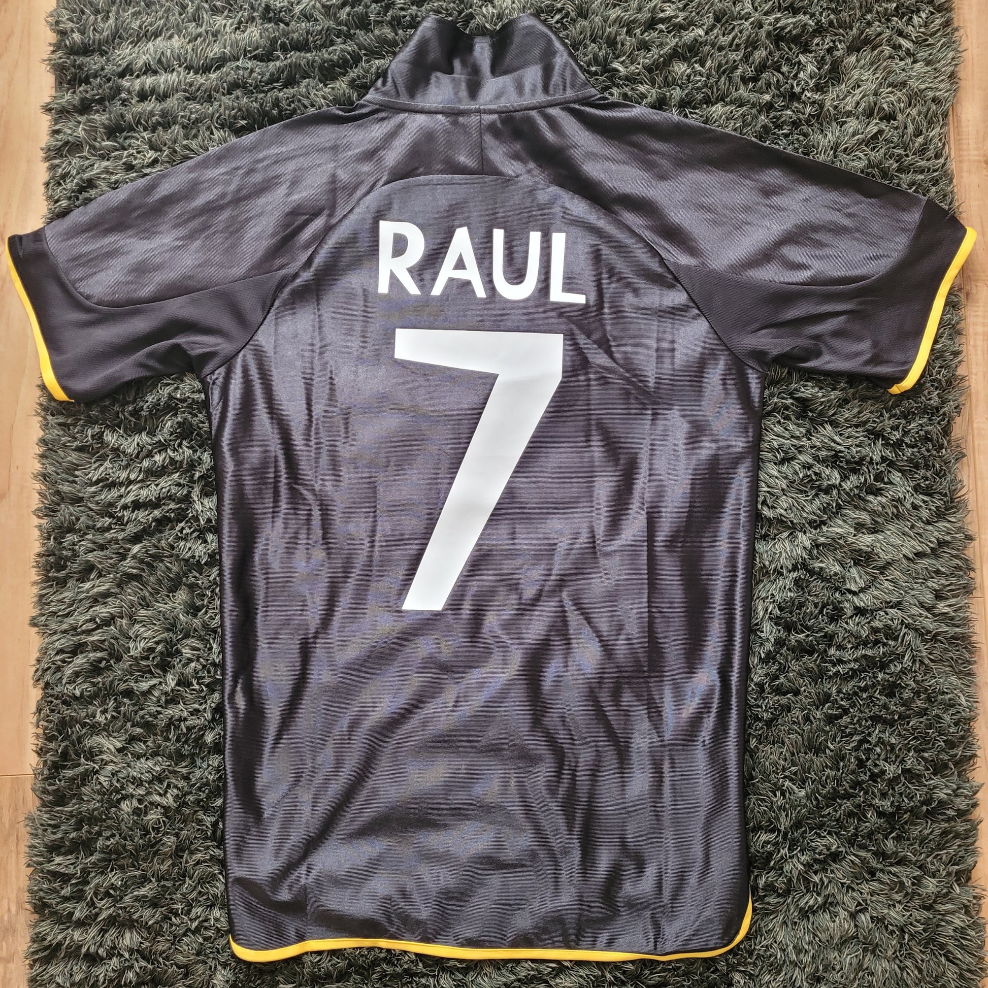 Rückansicht des Vintage Real Madrid Trikots Raul Saison 1999/2000 in Größe S, flach ausgelegt auf dunkelgrünem Teppich. Schwarzes Adidas-Trikot mit gelbem Saum zeigt den legendären Spielername "RAUL" und die Rückennummer 7 in Weiß – ein echtes Highlight für Sammler und Fans von Real Madrid Vintage Fußballtrikots