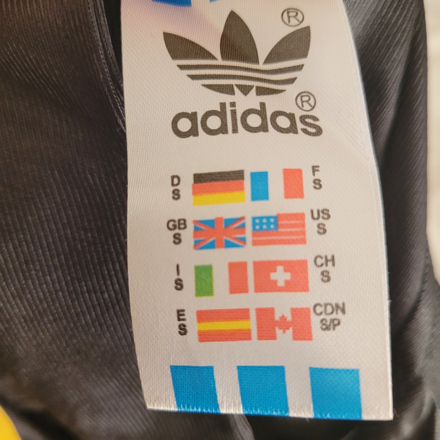 Detailaufnahme des Größenetiketts eines Vintage Real Madrid Trikots Raul Saison 1999/2000 von Adidas. Das Label zeigt internationale Größenangaben mit Länderflaggen (u. a. Deutschland, USA, Frankreich, Spanien) und ist am schwarzen Stoff des originalen Real Madrid Vintage Trikots befestigt – ideal zur Größenbestimmung für Sammler und Fans.