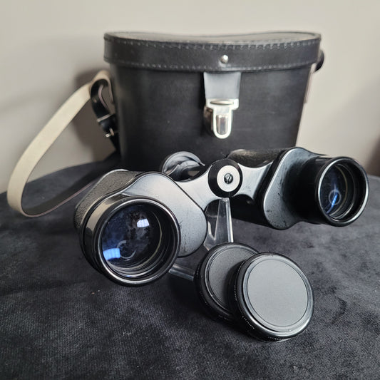 Ein Revue 8x30 Fernglas mit abnehmbaren Objektivabdeckungen auf einer dunklen Oberfläche. Im Hintergrund befindet sich eine schwarze Ledertasche mit silbernem Verschluss und beigem Trageriemen.