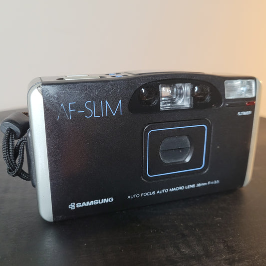Klassische Samsung AF-Slim Kompaktkamera mit 35mm f/3.5 Objektiv, eingebautem Blitz und schwarzem Design, auf einer Oberfläche liegend. Ideal für analoge Fotografie. 📸✨