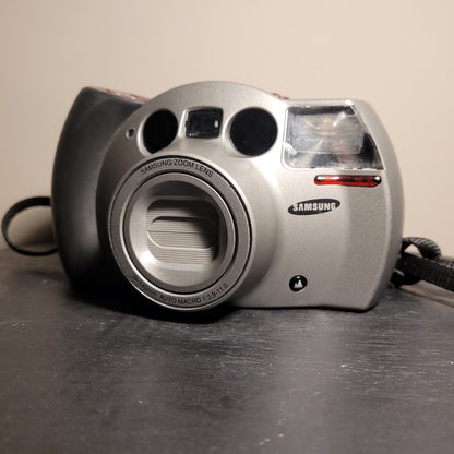Das Bild zeigt eine silberne Analogkamera der Marke Samsung. Die Kamera hat ein auffälliges Design mit einem großen Objektiv in der Mitte, das als 'Samsung Zoom Lens' gekennzeichnet ist. Um das Objektiv herum sind die Spezifikationen '38-140mm AUTO MACRO 1:3.9-11.0' zu sehen. Auf der rechten Seite der Kamera befindet sich ein Blitzlicht, und darunter ist das Samsung-Logo angebracht. Die Kamera hat auch einen Trageriemen, der auf der linken Seite sichtbar ist.