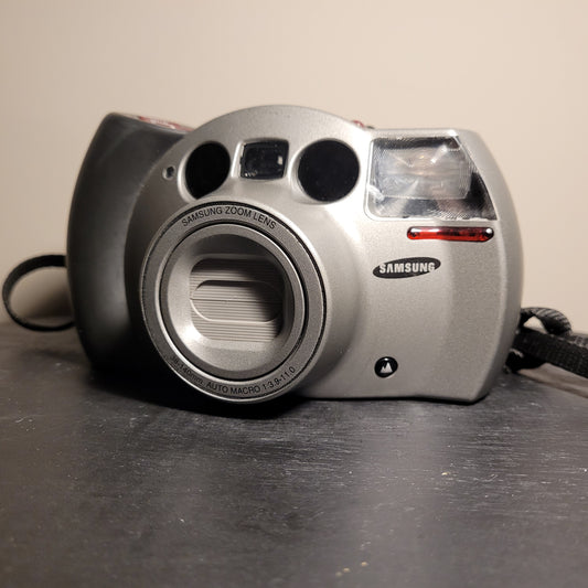 Das Bild zeigt eine silberne Analogkamera der Marke Samsung. Die Kamera hat ein auffälliges Design mit einem großen Objektiv in der Mitte, das als 'Samsung Zoom Lens' gekennzeichnet ist. Um das Objektiv herum sind die Spezifikationen '38-140mm AUTO MACRO 1:3.9-11.0' zu sehen. Auf der rechten Seite der Kamera befindet sich ein Blitzlicht, und darunter ist das Samsung-Logo angebracht. Die Kamera hat auch einen Trageriemen, der auf der linken Seite sichtbar ist.