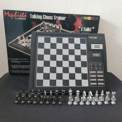 Das Bild zeigt das Schach Mephisto Talking Chess Trainer Modul von Saitek mit vollständigem Zubehör in seiner Originalverpackung (OVP). Das Schachgerät liegt auf einem schwarzen Untergrund mit weißem Hintergrund, wodurch die Details der Verpackung und des Moduls gut sichtbar sind. Neben dem Schachcomputer sind die Schachfiguren erkennbar, was die Vollständigkeit des Sets unterstreicht.