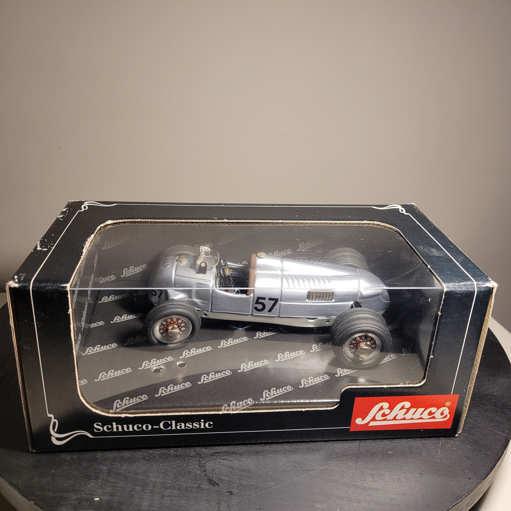 Das Bild zeigt ein Modellauto in einer Verpackung. Das Modellauto ist ein silberner Rennwagen mit der Nummer 57 auf der Seite. Die Verpackung ist schwarz mit einem transparenten Fenster, durch das das Auto sichtbar ist. Auf der Verpackung steht 'Schuco-Classic' und das Logo der Marke Schuco ist in der unteren rechten Ecke zu sehen.