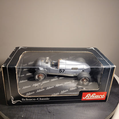 Das Bild zeigt ein Modellauto in einer Verpackung. Das Modellauto ist ein silberner Rennwagen mit der Nummer 57 auf der Seite. Die Verpackung ist schwarz mit einem transparenten Fenster, durch das das Auto sichtbar ist. Auf der Verpackung steht 'Schuco-Classic' und das Logo der Marke Schuco ist in der unteren rechten Ecke zu sehen.