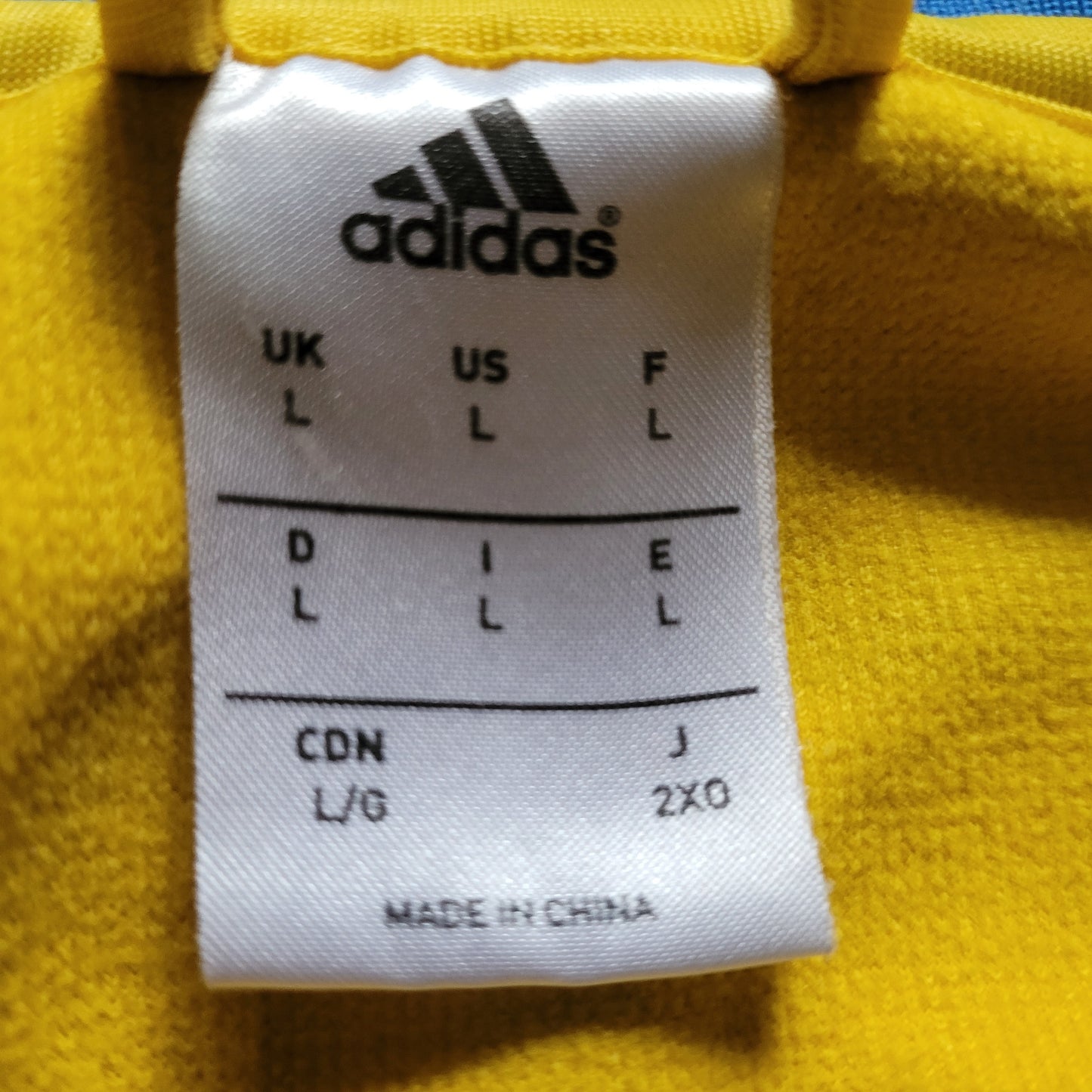 Größenetikett eines gelb-blauen Adidas Vintage Schweden Trainingsanzugs; weißes Label mit schwarzer Schrift zeigt internationale Größenangaben (u. a. UK L, US L, J 2XO) sowie „Made in China“ – eingenäht in gelben Stoff.