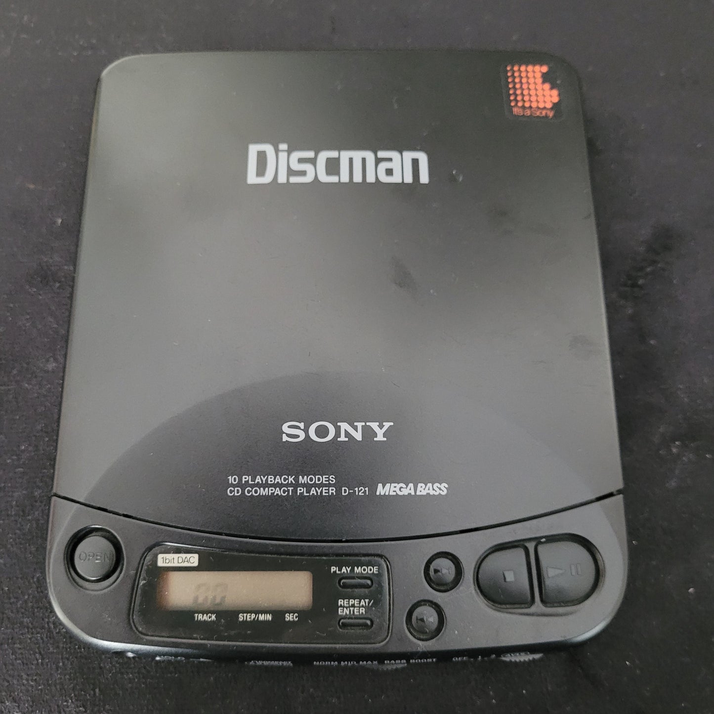 Sony Vintage Discman Mega Bass Model D-121 tragbarer CD-Player in Schwarz mit sichtbarem Sony-Logo und Discman-Schriftzug auf der Oberseite. Das Gerät zeigt die Aufschrift „10 Playback Modes“, „CD Compact Player D-121“ und „Mega Bass“. Rechts befinden sich Bedienelemente wie Play/Pause, Stop und Skip sowie ein digitales Display zur Wiedergabeanzeige. Der Sony Discman D-121 ist ein ikonisches Vintage-Audiogerät aus den 1990er Jahren mit klassischem Design