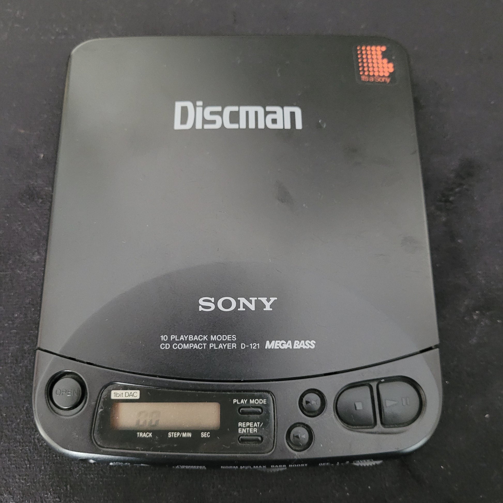 Sony Vintage Discman Mega Bass Model D-121 tragbarer CD-Player in Schwarz mit sichtbarem Sony-Logo und Discman-Schriftzug auf der Oberseite. Das Gerät zeigt die Aufschrift „10 Playback Modes“, „CD Compact Player D-121“ und „Mega Bass“. Rechts befinden sich Bedienelemente wie Play/Pause, Stop und Skip sowie ein digitales Display zur Wiedergabeanzeige. Der Sony Discman D-121 ist ein ikonisches Vintage-Audiogerät aus den 1990er Jahren mit klassischem Design