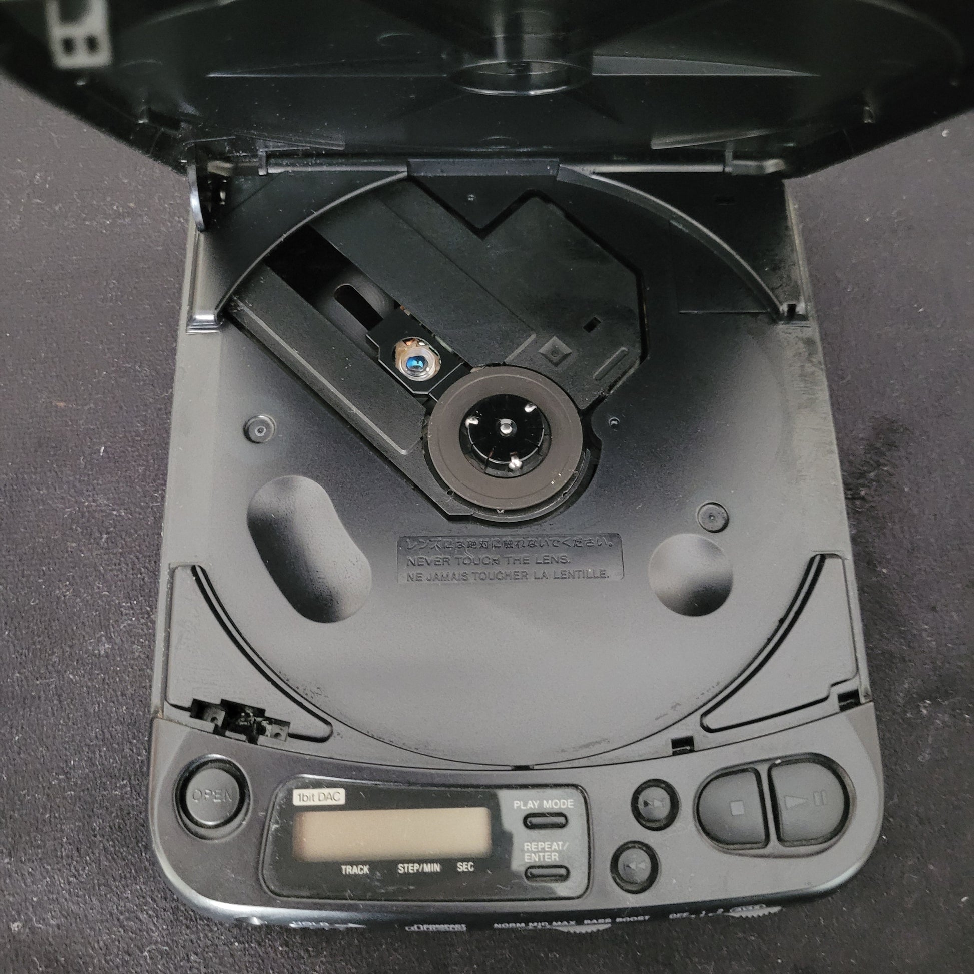 Geöffnetes Sony Vintage Discman Mega Bass Model D-121 mit sichtbarem CD-Laufwerk, Laserlinse und Warnhinweis „NEVER TOUCH LENS / NE JAMAIS TOUCHER LA LENTILLE“. Rechts im Bild befinden sich Bedientasten wie Play Mode, Repeat, Step/AM sowie ein digitales Display zur Anzeige der Wiedergabeinformationen. Das Innenleben des tragbaren CD-Players zeigt die klassische Technik der 1990er Jahre und unterstreicht den authentischen Vintage-Charakter des Sony Discman D-121.