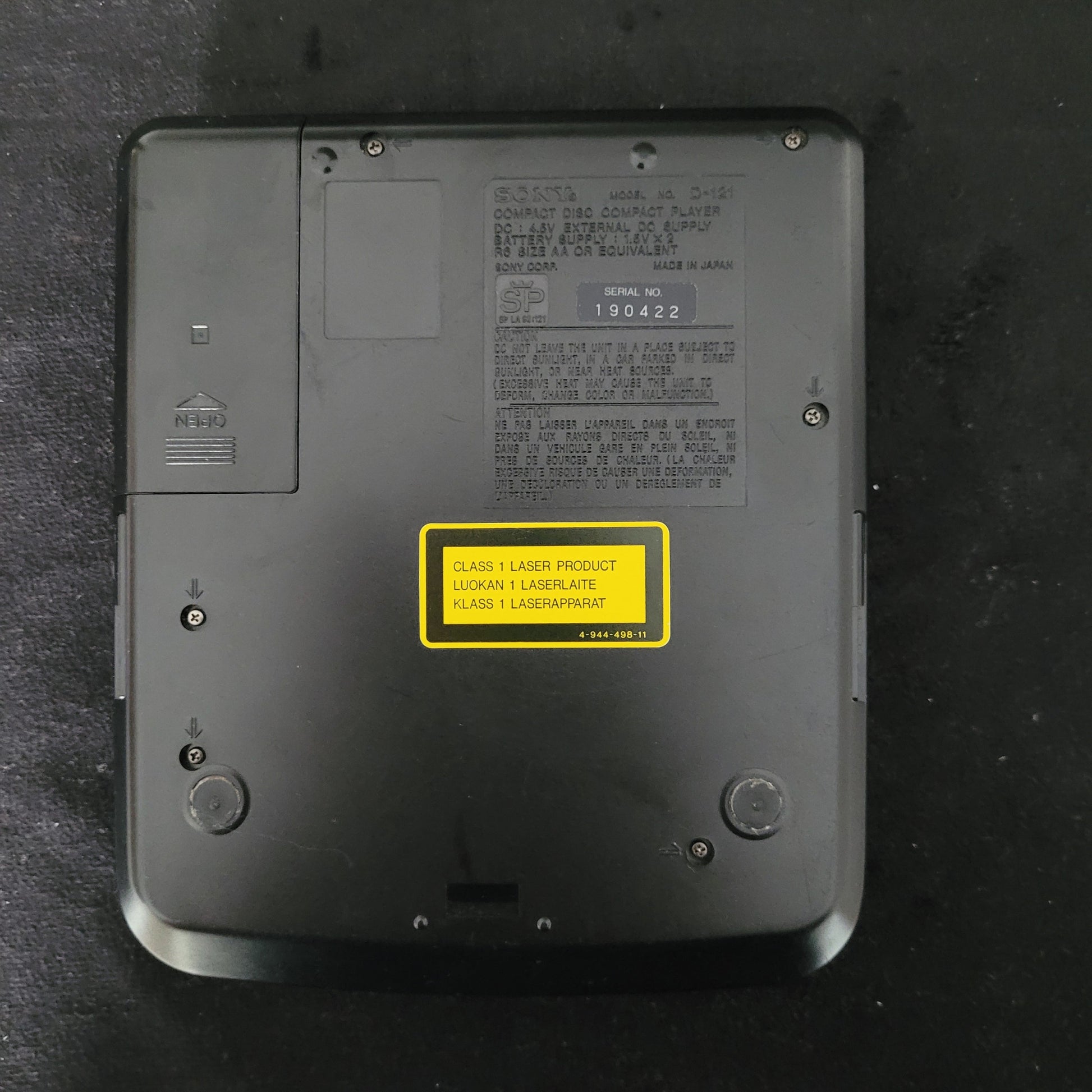 Unterseite des Sony Vintage Discman Mega Bass Model D-121 mit sichtbarem Batteriefach, mehreren Schrauben und technischen Etiketten. Zu sehen sind ein gelbes Warnschild mit der Aufschrift „CLASS 1 LASER PRODUCT / LUOKAN 1 LASERLAITE / KLASS 1 LASERAPPARAT“ sowie ein graues Typenschild mit Modellnummer, Seriennummer und Herstellerangaben. Das robuste Gehäuse des tragbaren CD-Players unterstreicht den authentischen Vintage-Charakter und zeigt die hochwertige Verarbeitung typischer Sony-Geräte der 1990er Jahre
