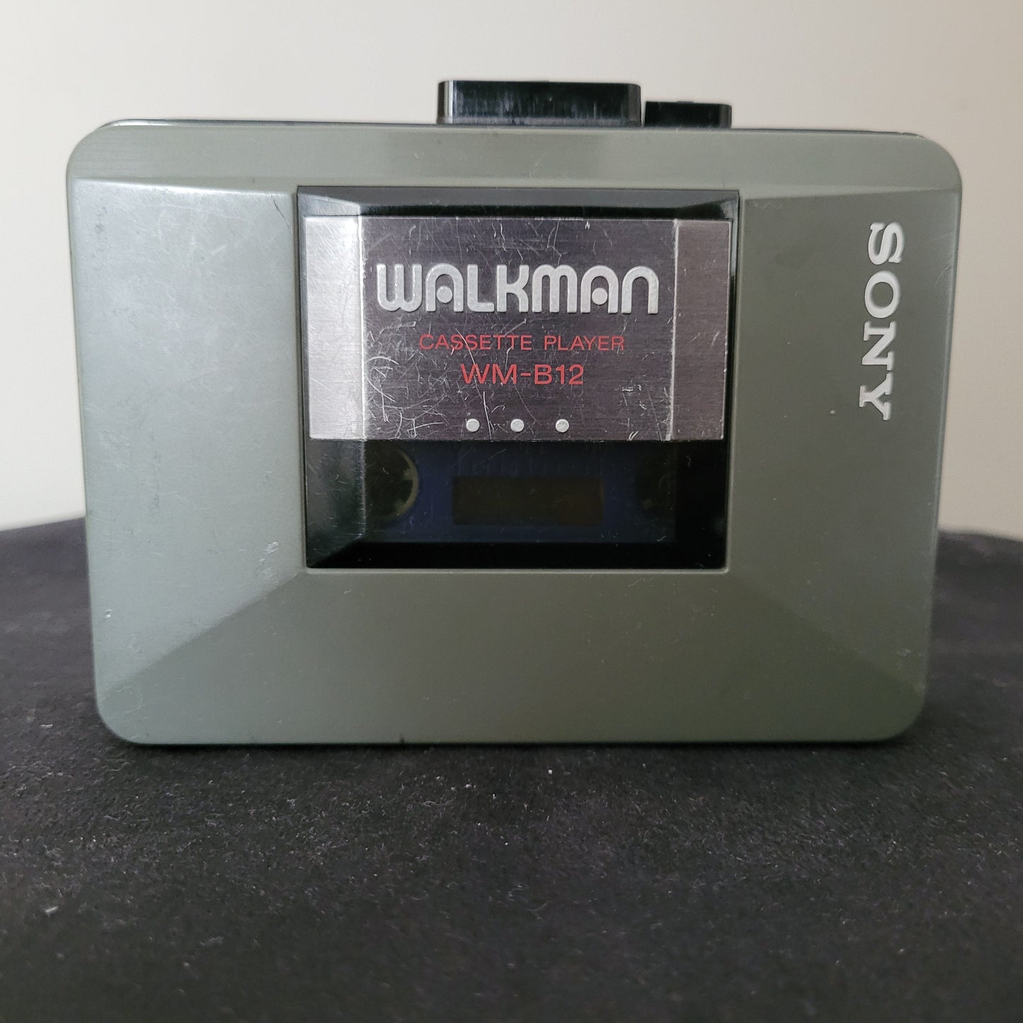 Sony Walkman WM-B12 Vintage Kassettenspieler in graugrünem Gehäuse mit Sichtfenster für Kassette, sichtbarem "WALKMAN CASSETTE PLAYER WM-B12"-Label und seitlich angebrachtem silbernen "SONY"-Logo; klassisches tragbares Audiogerät aus den 1980er Jahren, ideal für Retro-Technikliebhaber und Sammler von nostalgischer Unterhaltungselektronik.