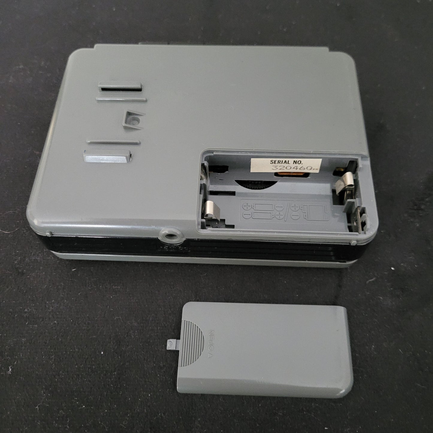 Rückansicht eines geöffneten Sony Walkman WM-B12 Kassettenspielers in graugrünem Gehäuse mit freiliegendem Batteriefach, sichtbaren Metallkontakten und eingeprägten Polaritätssymbolen zur korrekten Batterieausrichtung; rechts daneben liegt die abgenommene Batteriefachabdeckung. Im Inneren befindet sich ein Etikett mit der Seriennummer "SERIAL NO. 3204604". Das Gerät zeigt Schrauböffnungen und Befestigungsschlitze – ideal für Technikliebhaber und Sammler von Vintage-Audioelektronik.