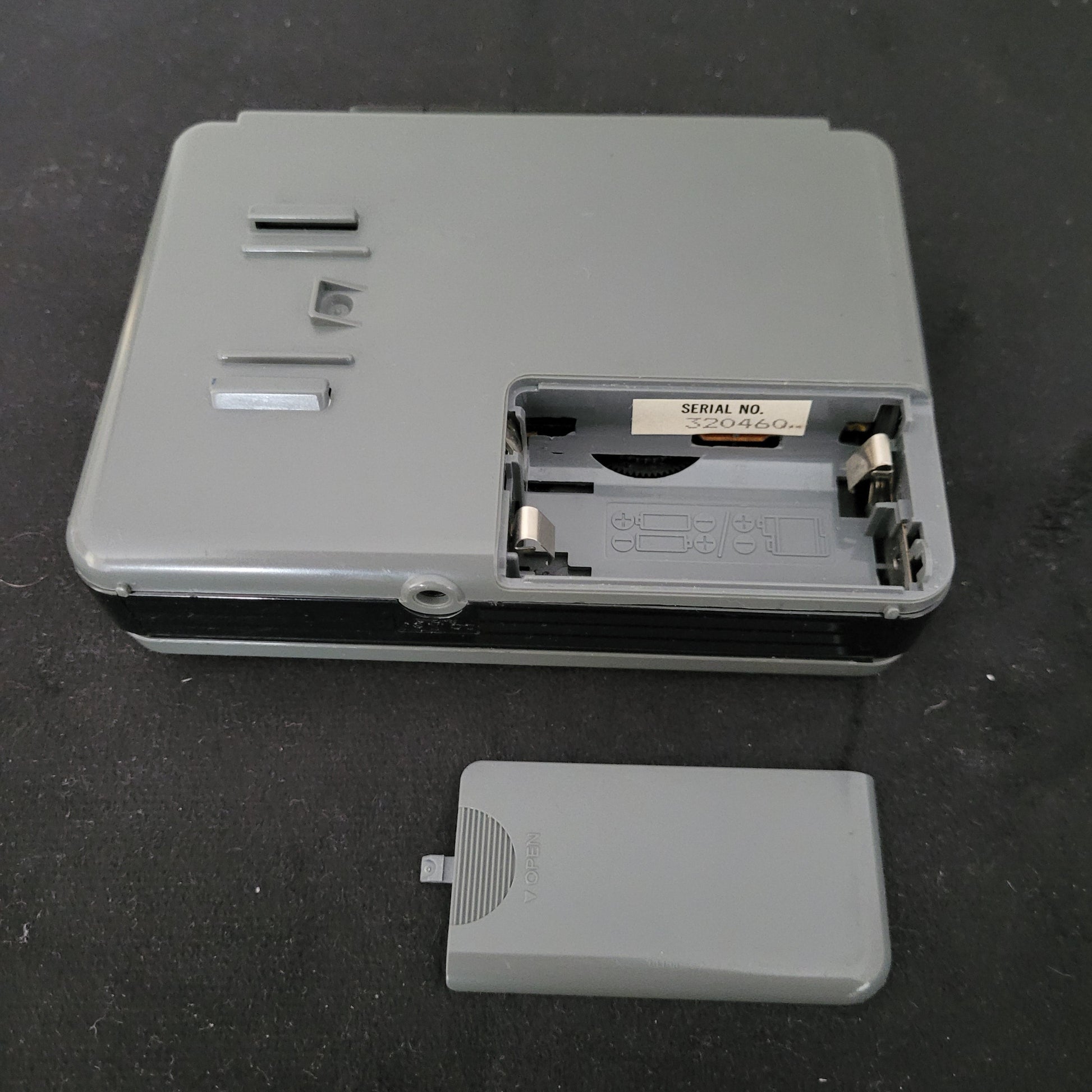 Rückansicht eines geöffneten Sony Walkman WM-B12 Kassettenspielers in graugrünem Gehäuse mit freiliegendem Batteriefach, sichtbaren Metallkontakten und eingeprägten Polaritätssymbolen zur korrekten Batterieausrichtung; rechts daneben liegt die abgenommene Batteriefachabdeckung. Im Inneren befindet sich ein Etikett mit der Seriennummer "SERIAL NO. 3204604". Das Gerät zeigt Schrauböffnungen und Befestigungsschlitze – ideal für Technikliebhaber und Sammler von Vintage-Audioelektronik.