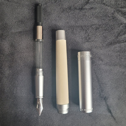 Das Bild zeigt die Teile eines hochwertigen Füllfederhalters auf einem dunklen Stoff: Metallfeder mit Tintenleiter und Tintenreservoir, zylindrischer Schaft aus hellbeigem Material und die Metallkappe mit den Aufschriften 'STAEDTLER' und 'MADE IN GERMANY