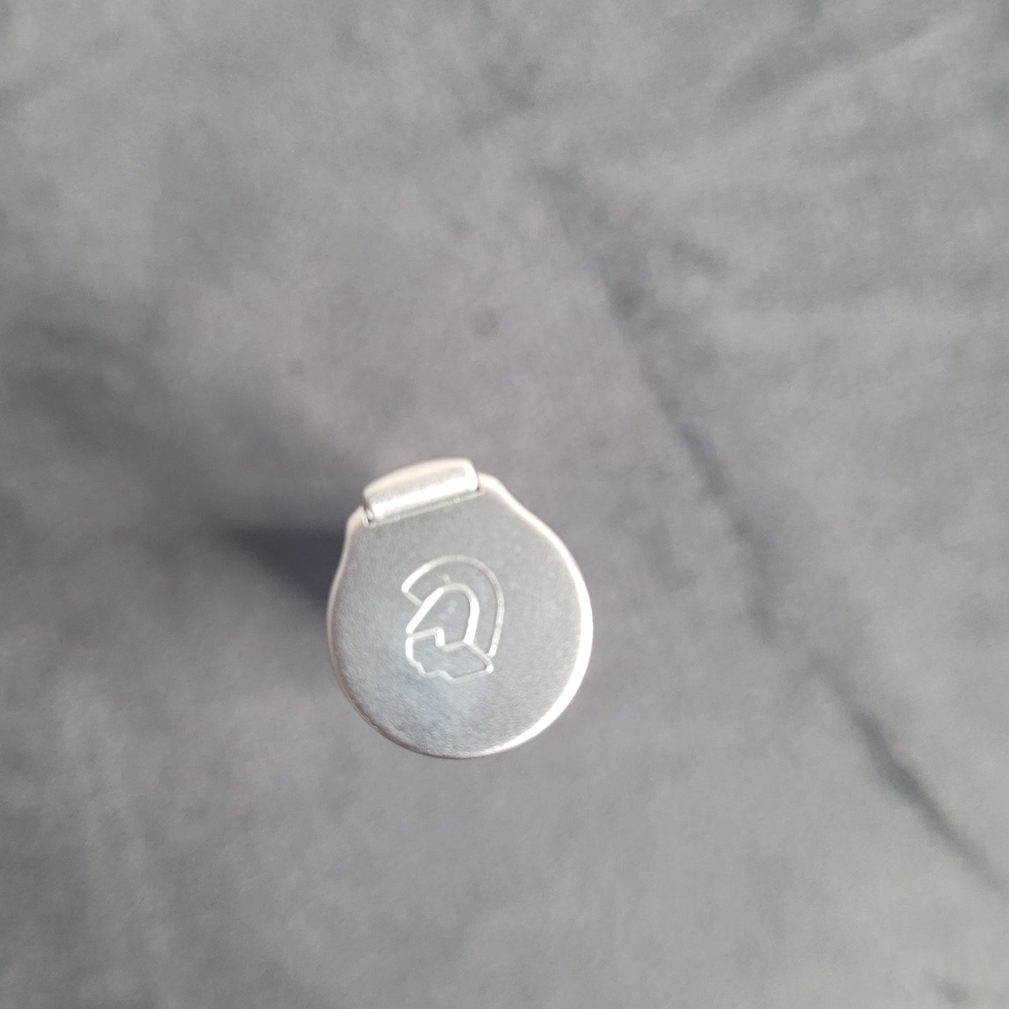 Das Bild zeigt die Draufsicht eines metallischen Objekts mit einem runden Deckel. Der Deckel ist mit einem gravierten Logo oder Symbol verziert, das wie ein stilisiertes 'G' oder ein ähnliches Design aussieht. Das Objekt liegt auf einem leicht texturierten, einfarbigen Hintergrund.