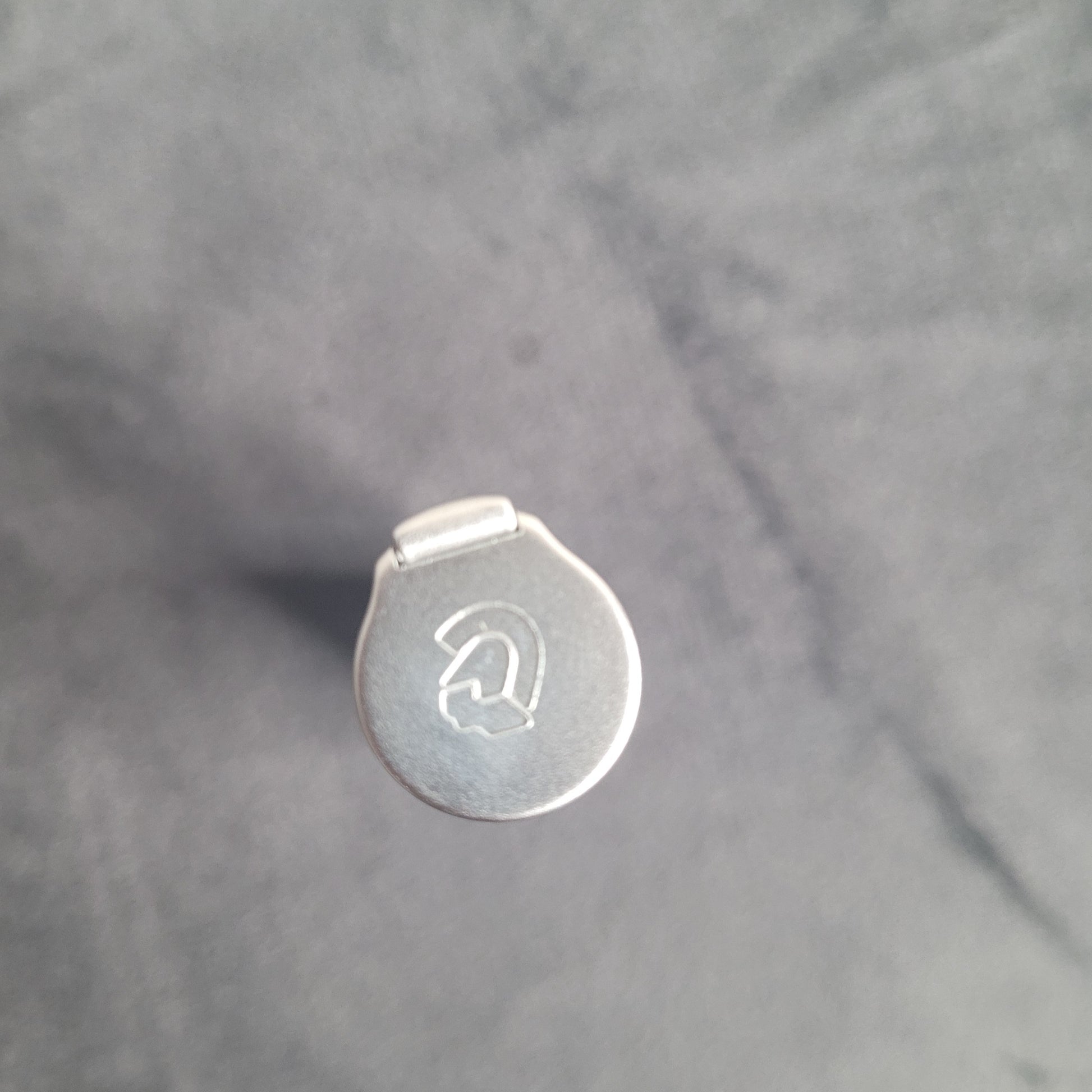 Das Bild zeigt die Draufsicht eines metallischen Objekts mit einem runden Deckel. Der Deckel ist mit einem gravierten Logo oder Symbol verziert, das wie ein stilisiertes 'G' oder ein ähnliches Design aussieht. Das Objekt liegt auf einem leicht texturierten, einfarbigen Hintergrund.