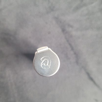 Das Bild zeigt die Draufsicht eines metallischen Objekts mit einem runden Deckel. Der Deckel ist mit einem gravierten Logo oder Symbol verziert, das wie ein stilisiertes 'G' oder ein ähnliches Design aussieht. Das Objekt liegt auf einem leicht texturierten, einfarbigen Hintergrund.