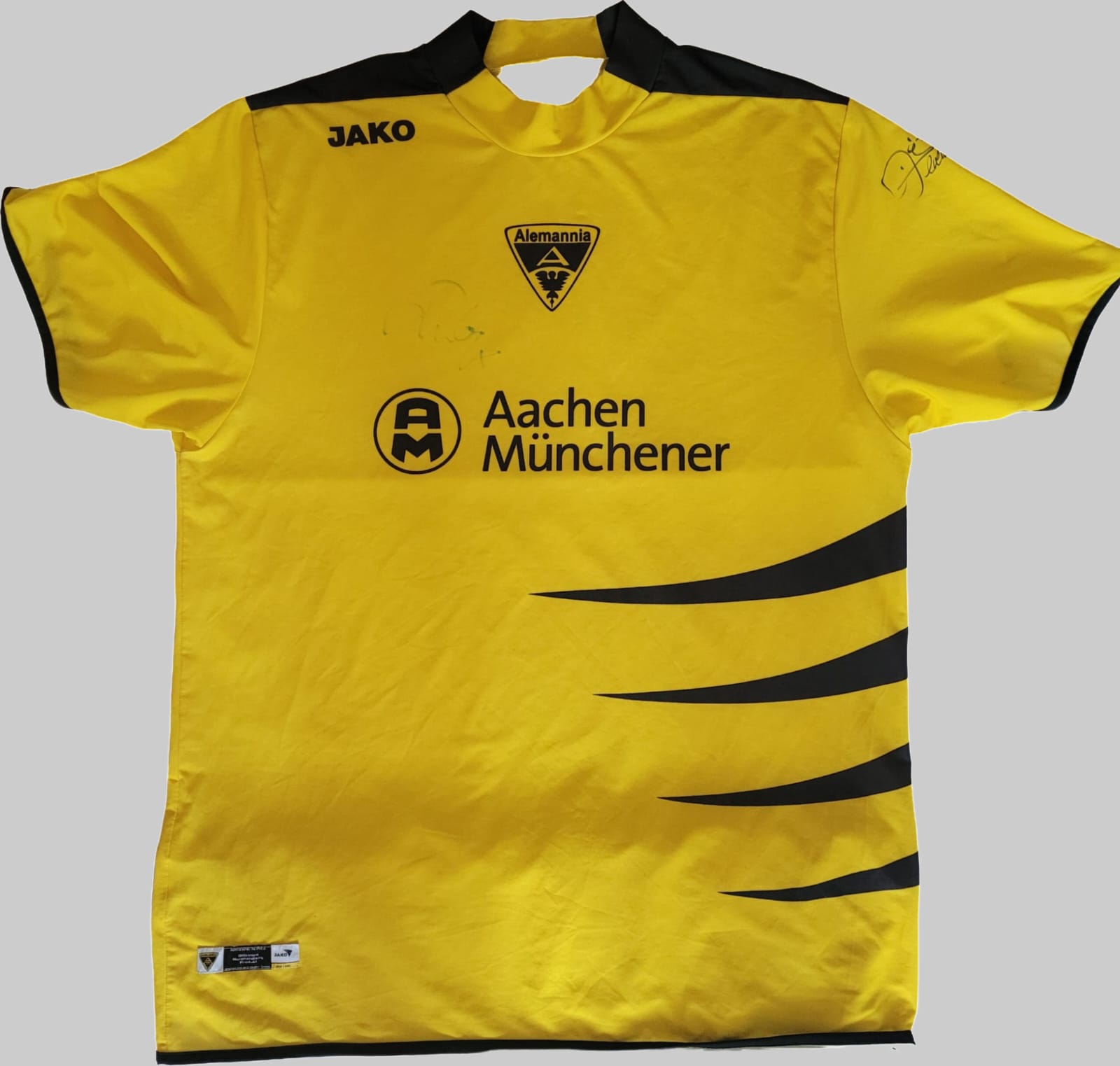 TSV Alemannia Aachen Vintage Trikot 2006/2007 – Vorderseite mit Original Signatur Das Bild zeigt die Vorderseite des TSV Alemannia Aachen Vintage Trikots 2006/2007, platziert auf einem grauen Hintergrund. Das Trikot ist gelb mit schwarzen Akzenten an Schultern und Seiten. Auf der linken Brustseite befindet sich das JAKO-Logo, mittig prangt das Vereinswappen von Alemannia Aachen, darunter der Schriftzug „Aachen Münchener“ mit zugehörigem Logo. Eine Original Signatur ist auf der rechten Brustseite sichtbar.